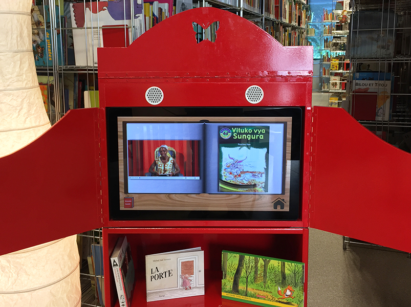 Photo du kiosque à histoires multilingues, volets ouverts et écran qui diffuse une histoire avec une lectrice et à côté d'elle les images du livre qui défilent